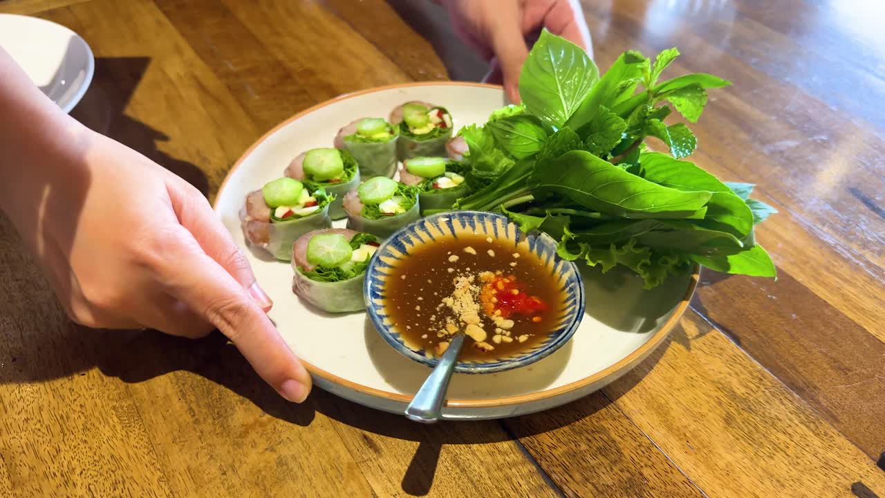 Hands arrange a Vietnamese dish with herbs and dipping sauce on a wooden table under natural light