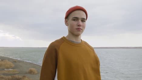 vista frontal de un adolescente con gorro naranja y sudadera posando en la cámara en la cima de la montaña en la playa en un día ventoso