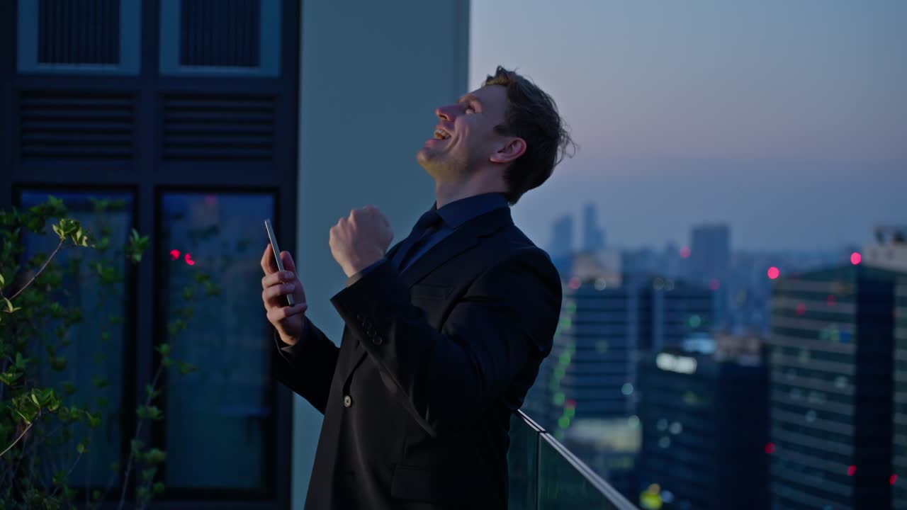 Joyful businessman celebrating success after receiving good news on smartphone at rooftop terrace during sunset - slow reveal