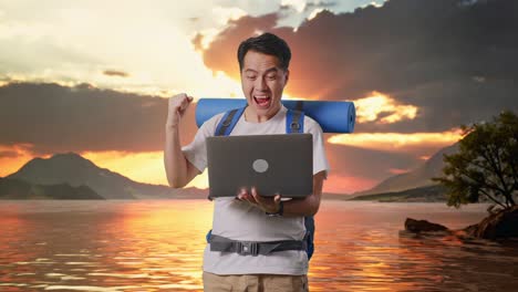excursionista masculino asiático con mochila de montañismo mirando una computadora portátil y gritando meta celebrando el éxito en un lago