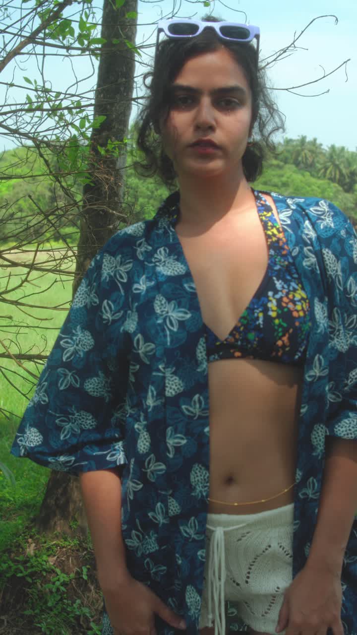 A close-up of a woman in a bikini top and blue kaftan, adjusting her top while standing among trees in a tropical forest. Capturing summer confidence, beach fashion, and natural beauty