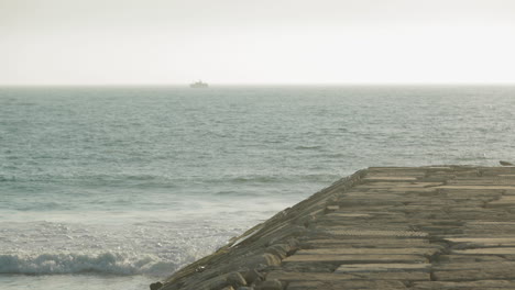 Distant-View-From-Seawall-Of-A-Boat-Sailing-Across-The-Ocean-With-Waves-Splashing