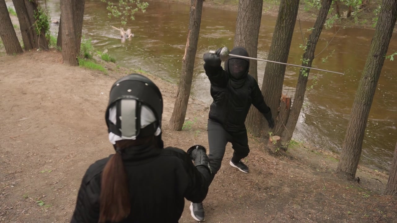 Two people in black fencing gear engage with swords near riverbank demonstrating controlled movements and techniques. scene features natural outdoor elements like trees and water providing