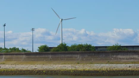 a large wind turbine spins steadily beside a coastal barrier under clear daylight in dundee, scotland, with static camera and calm atmosphere