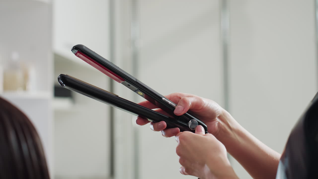 코카서스인 스타일리스트가 거울 근처에서 빛나는 헤어 스트레이트너를 열고 있어요. 미용실 선반에는 병들이 보이고, 머리를 다듬기 전에 정확하게 잡고 장치를 점검하는 모습은 전문적인 준비 장면을 연출합니다.