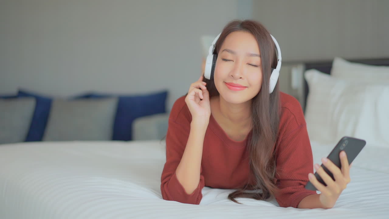 행복한 예쁜 아시아 여성이 캐주얼한 옷을 입고 호텔 침대에 누워 전면을 보며 전화를 보고 음악을 듣습니다.