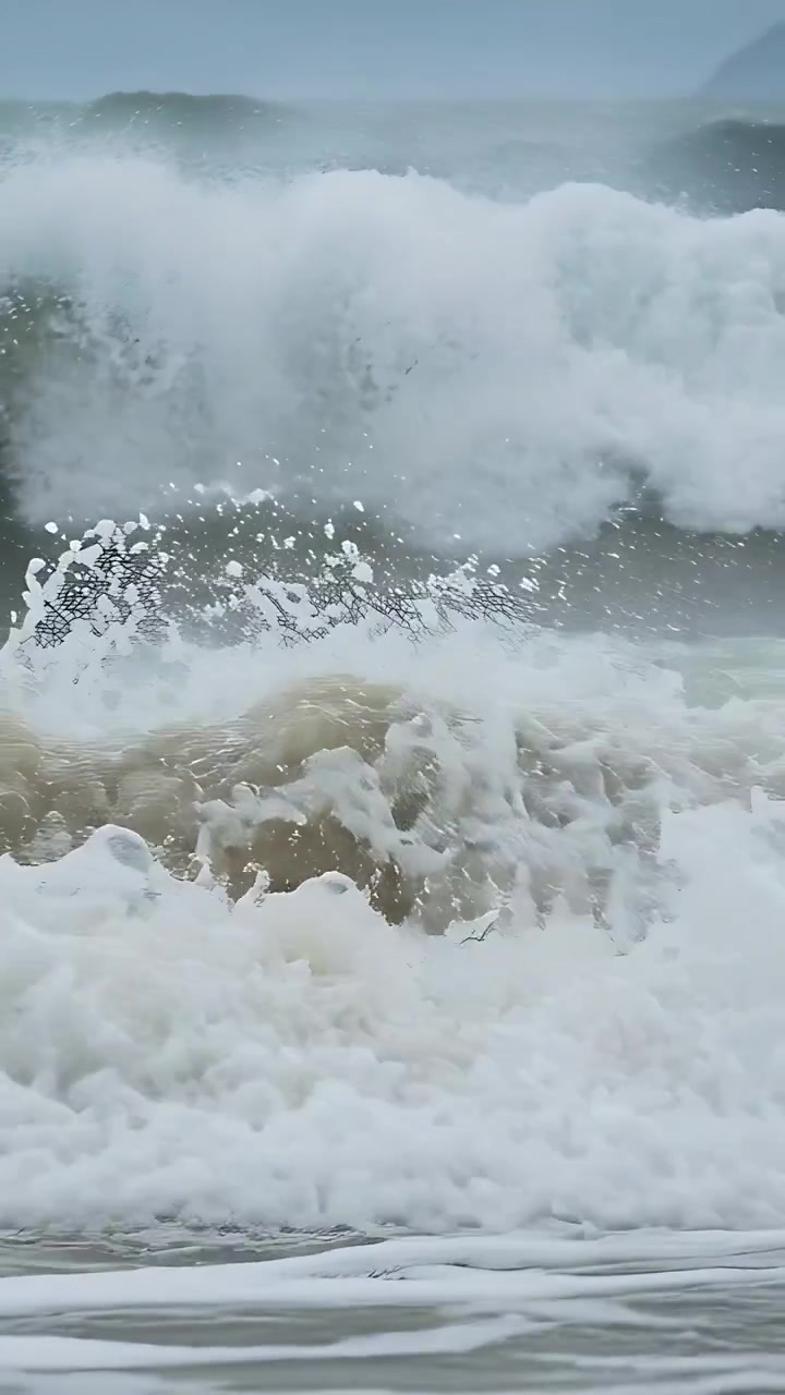 Video verticale: Onde oceaniche impetuose che si infrangono, spingendo schiuma bianca sulla sabbia bagnata, mosse da una mareggiata crescente.