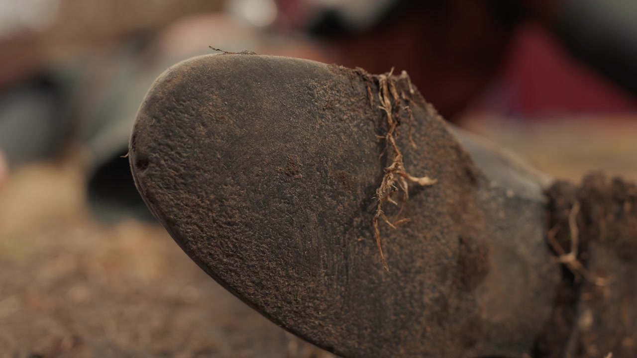 Low-angle close-up shows worn boot toe pressing into rough ground, with heavy scuff marks, lifted frayed stitching, dusty leather, and shallow depth of field for gritty tactile detail