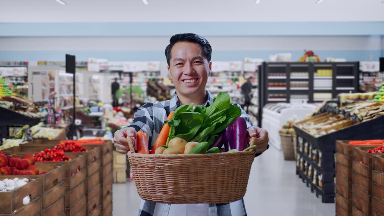 Agricultor asiático sujetando una cesta con verduras y sonriendo amigablemente en un supermercado