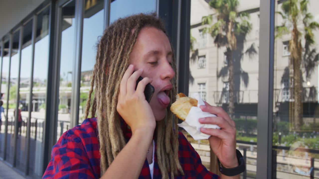 hombre de raza mixta con dreadlocks caminando por la calle comiendo sándwich y hablando en el teléfono inteligente
