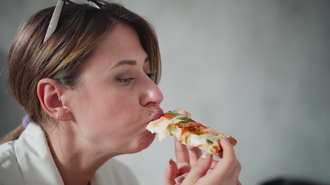 Close up of young woman with tied hair and eyeglasses on head joyfully biting cheesy pizza slice during break, smiling slightly while savoring delicious meal in relaxed home office atmosphere