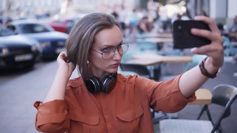 A-stylish-girl-in-the-middle-of-the-street-takes-a-photo-on-her-cell-phone,-fixing-her-hair,-looks-for-and-angle.-Headphones-on