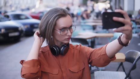 una chica elegante en el medio de la calle toma una foto en su teléfono celular, arreglando su cabello, busca y ángulo. auriculares en