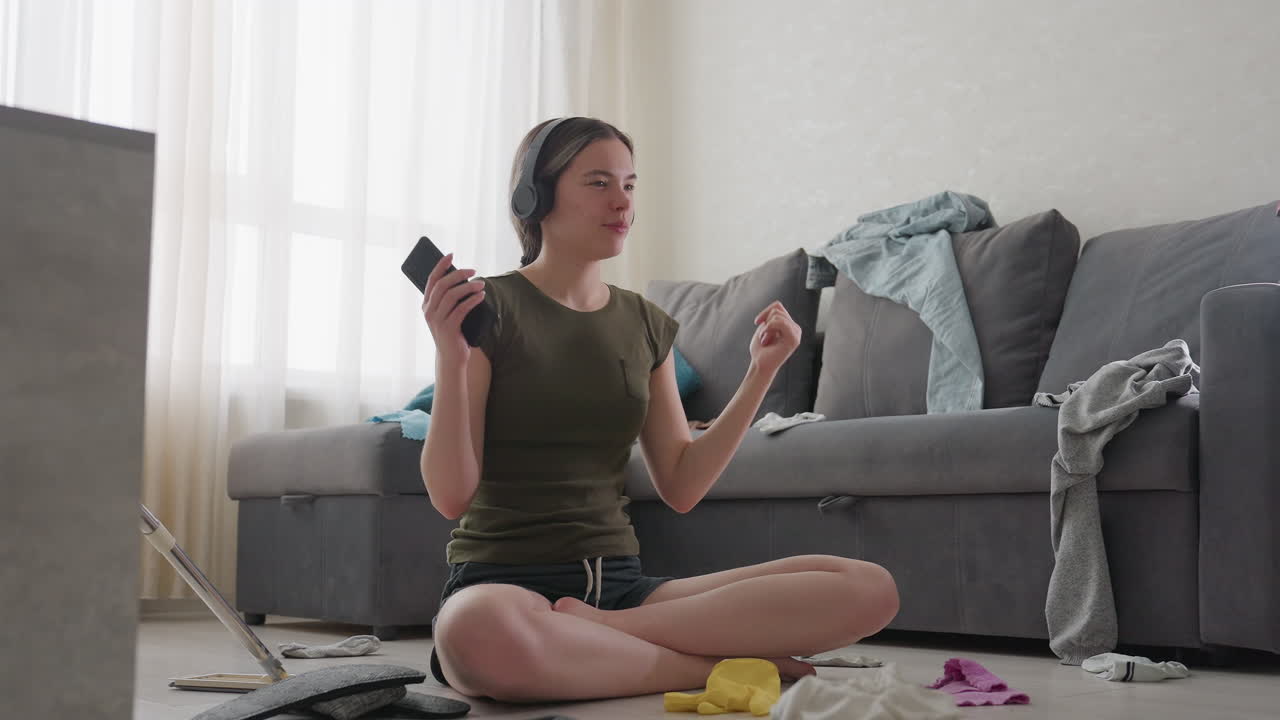 Student seated cross legged indoors dancing joyfully with phone in hand while listening to music on headphones, casual shorts and t shirt, scattered clothes on sofa in background