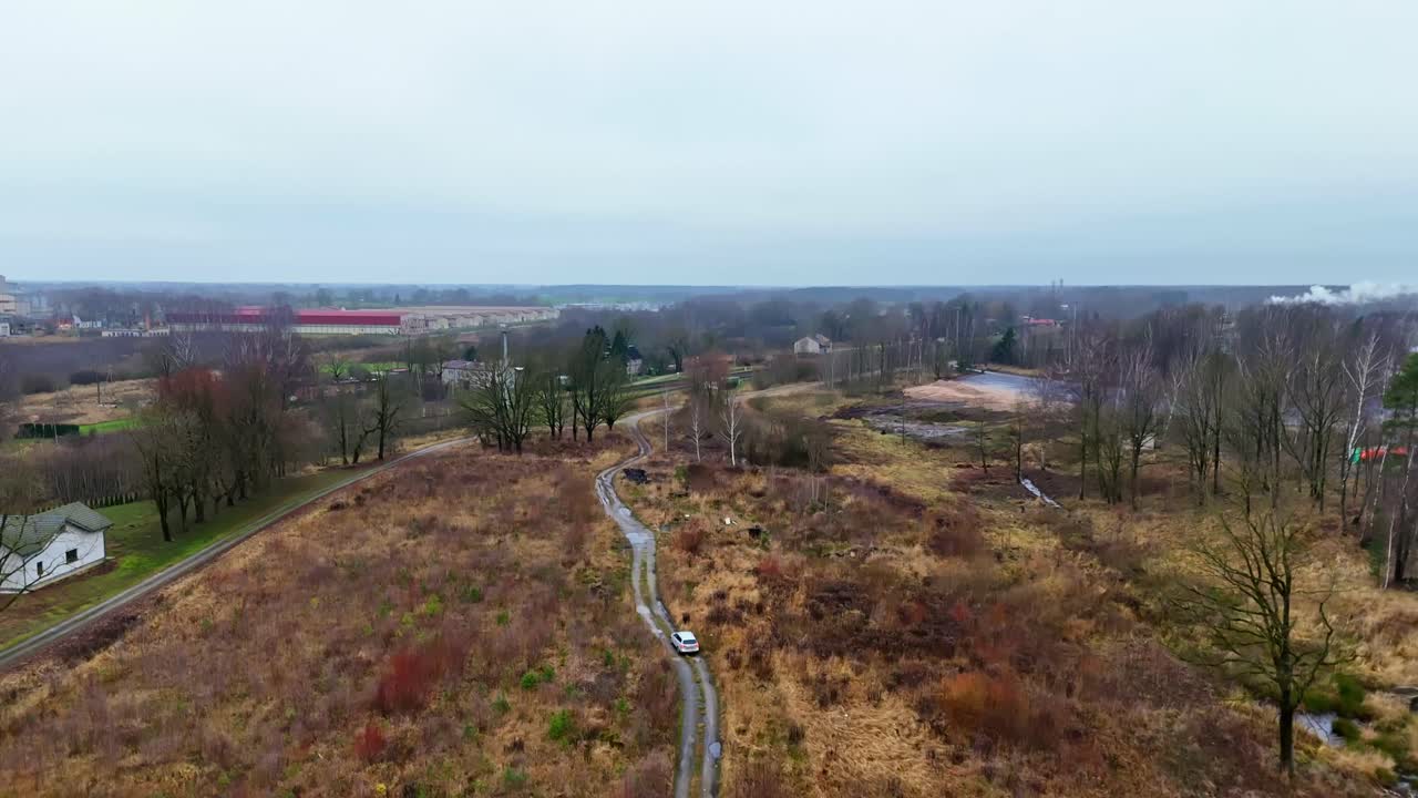 Ampia ripresa aerea di una zona industriale di campagna con linee ferroviarie, piccole fabbriche che emettono vapore e una strada rurale che attraversa erba secca e alberi nel tardo autunno