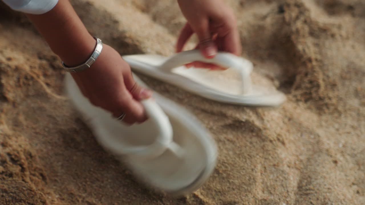 Hvite sandaler ligger på sandstrand, person henter paret med ømme hender, nærbilde av teksturert sand og falmende fotspor, varmt gyllent lys, rolig nostalgisk stemning og stille tilfluktssted ved kysten.