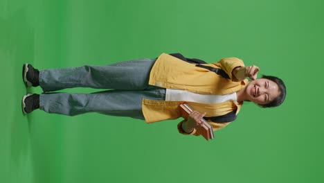 full body of asian woman student with a backpack and some books touching her chest then smiling and pointing to camera being proud of herself in the green screen background studio