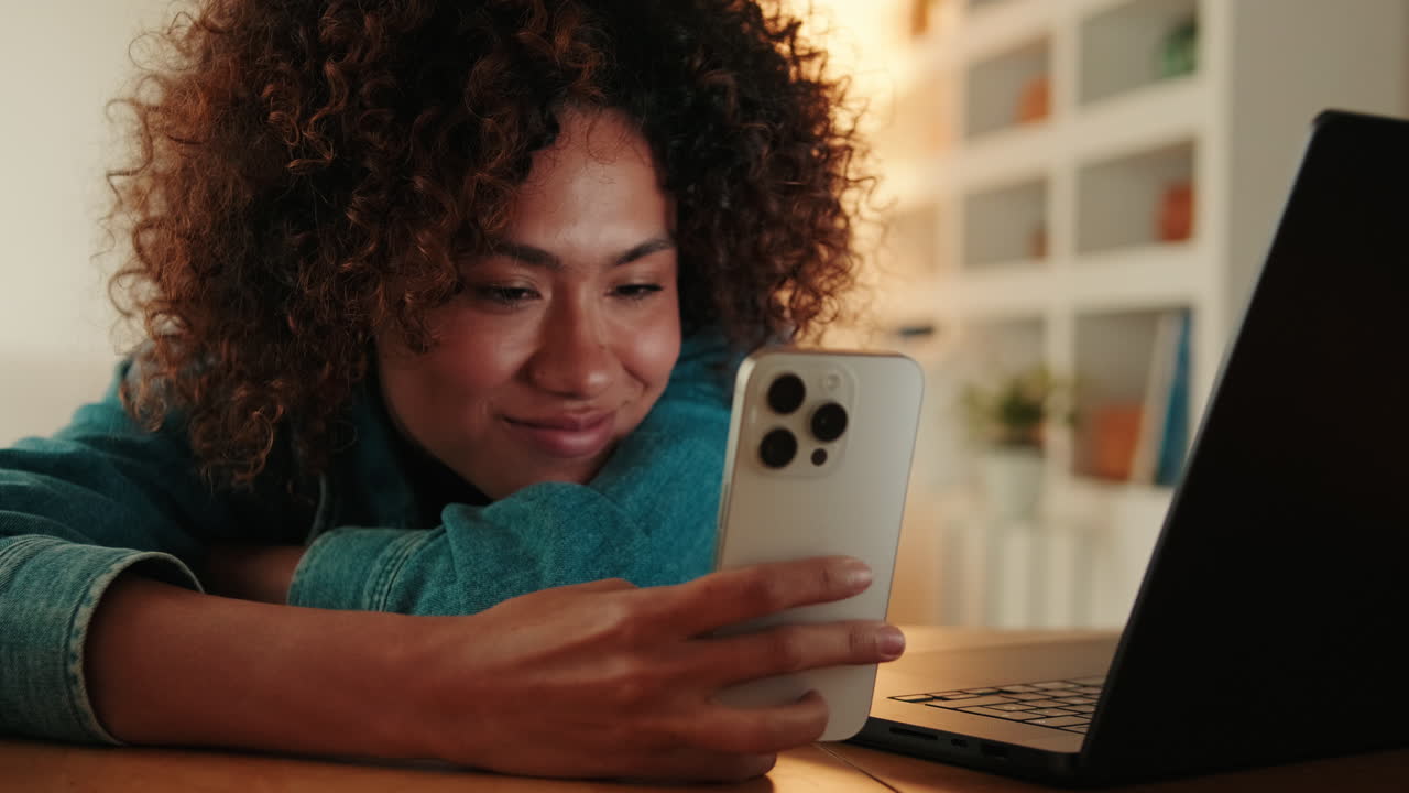 Young Woman Smiling While Looking at Smartphone Next to Laptop