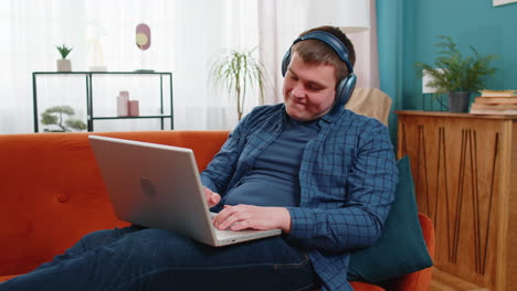 happy relaxed man in wireless headphones sitting on home couch listening music in laptop netbook
