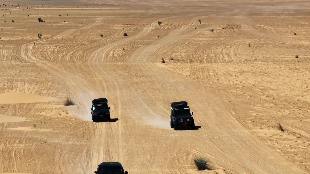 Ripresa cinematografica con drone di un convoglio overland fuoristrada che attraversa le vaste e desolate pianure sabbiose del deserto del Sahara tunisino sotto un cielo luminoso.