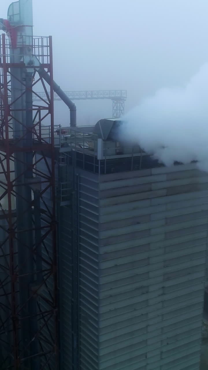 Grain processing plant premises on gray foggy day. Functioning enterprise producing heavy smoke into air. Aerial view. Vertical video
