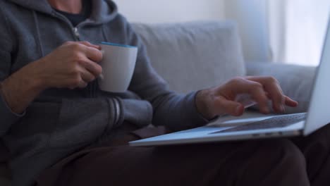 Young-man-drinks-from-a-mug-while-typing-on-laptop-at-home
