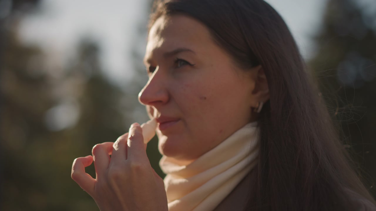 Primo piano di donna che tiene ed esamina un oggetto nella sua mano all'aperto con alberi visibili. La donna indossa una sciarpa di colore chiaro, concentrandosi intensamente sul piccolo oggetto che tiene vicino al viso sotto una luce soffusa.