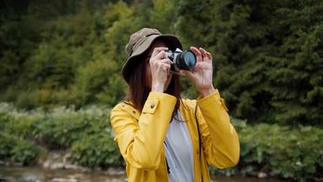 a brunette girl in a yellow hiking jacket photographs a forest and a mountain river with a camera