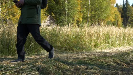 Adult-man-with-backpack-walks-past-old-barn-and-grain-field-on-hiking-trail,-yellow-autumn-colors,-camera-tracking