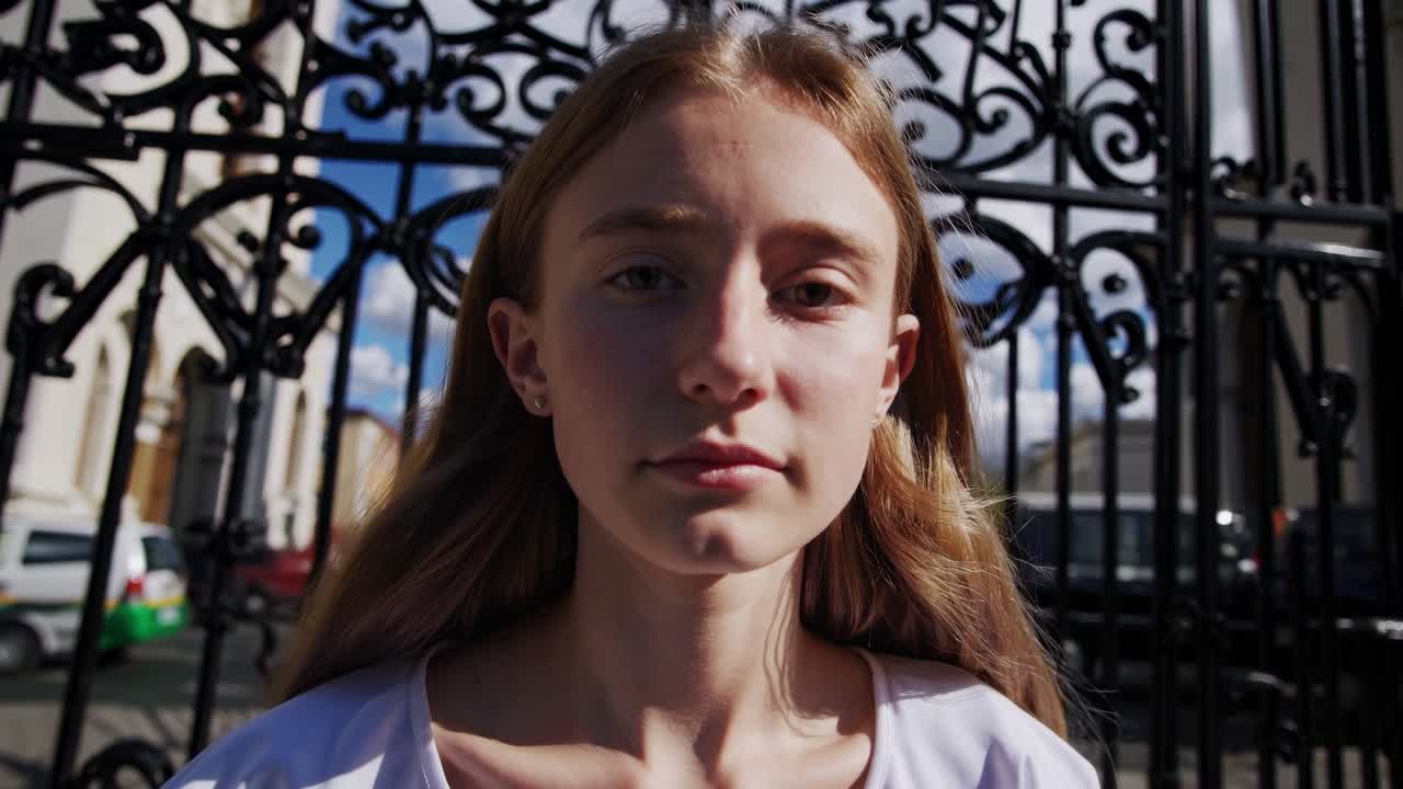 Portrait of a young blonde woman standing before an ornate metal gate, with a vibrant city and blue sky in the background, embodying urban life and youthful confidence