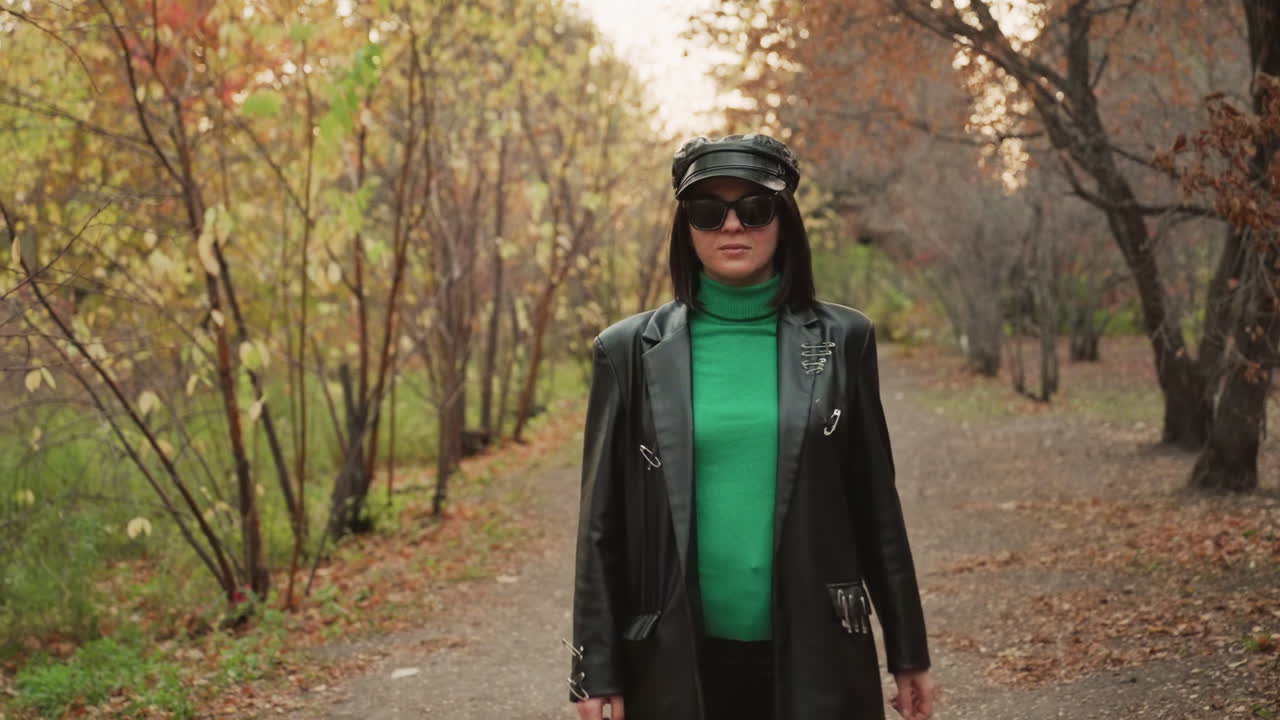 Mulher de jaqueta de couro caminhando por um parque outonal e frondoso, roqueira confiante caminhando em direção à câmera, gola alta verde e boné brilhante, óculos de sol escuros, tons quentes cinematográficos e folhas caídas, moda