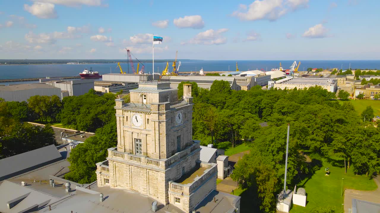 Aerial view orbiting around the Estonian flag fluttering top of Maritime Academy clock tower, old style building in Tallinn Kopli industrial district. Peninsula shipyard with cranes in the distance