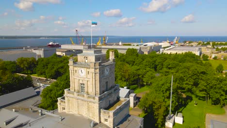 aerial view orbiting around the estonian flag fluttering top of maritime academy clock tower, old style building in tallinn kopli industrial district. peninsula shipyard with cranes in the distance