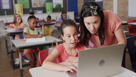 una feliz maestra diversa que enseña a las escolares usando una computadora portátil en el aula de la escuela primaria