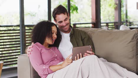 Sonriente-Pareja-De-Raza-Mixta-Teniendo-Una-Videollamada-En-Una-Tableta-Digital-En-El-Sofá-De-La-Casa-De-Vacaciones