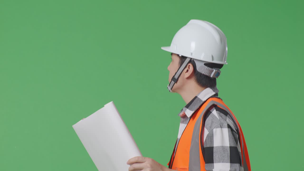 으로 가까이 쳐다보기: 안전 헬을 착용한 아시아 남성 엔지니어가 손에 청사진을 들고 녹색 스크린 배경 스튜디오에서 는 동안 주변을 둘러보고 있습니다.