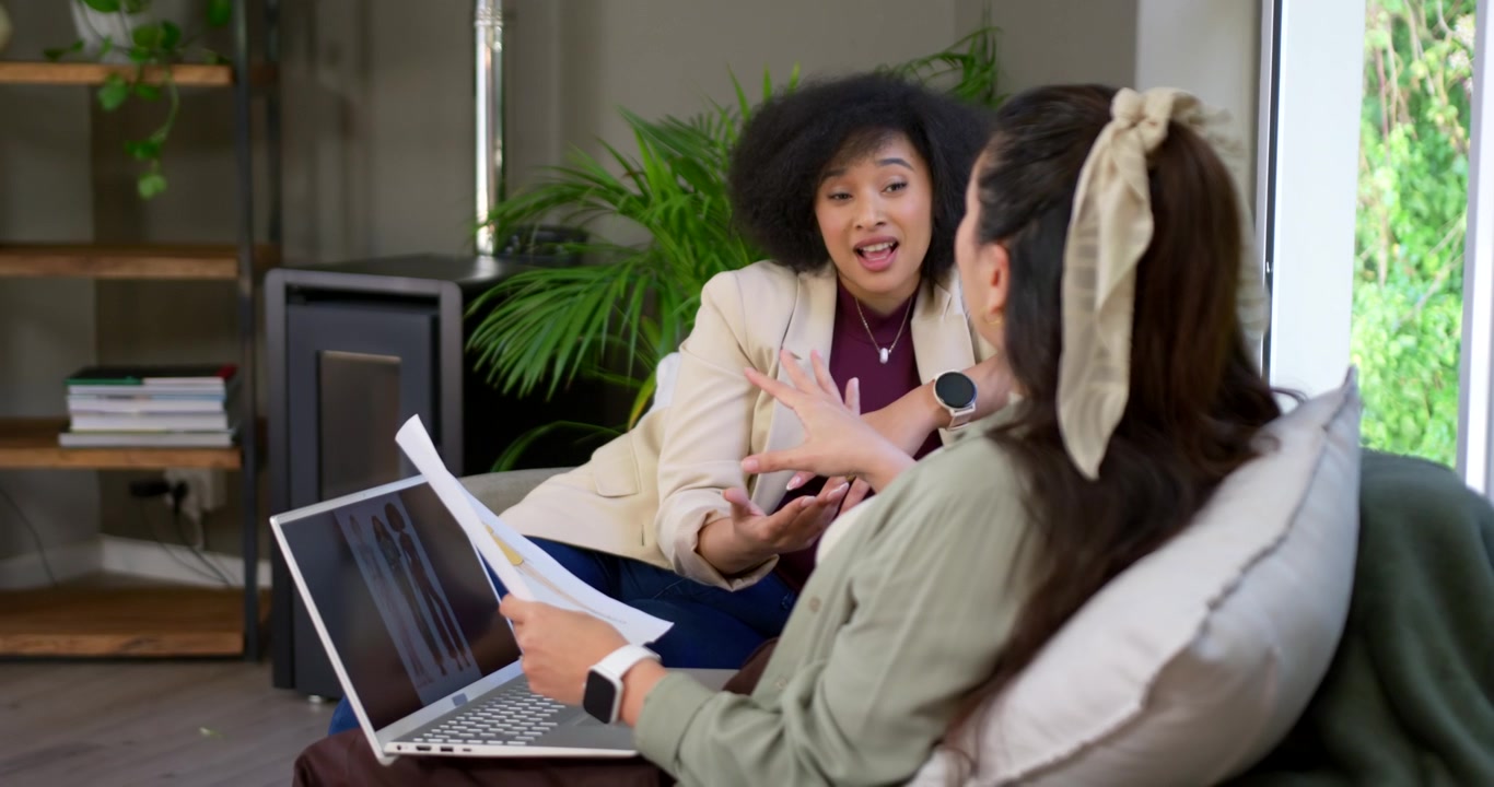 Diverse female colleagues prompted by printed documents reviewing and pointing at laptop on couch