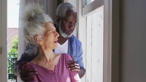 mixed race senior couple talking to each other while looking out of the window at home