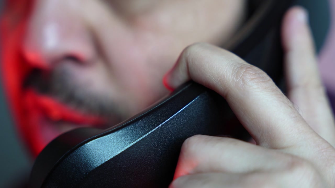 Un primer plano extremo de un hombre sosteniendo un auricular de teléfono fijo negro en su oído. La escena presenta una dramática iluminación de estudio roja y azul con poca profundidad de campo que desenfoca su rostro