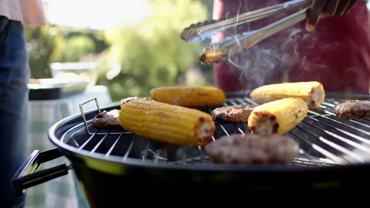 Grilling corn and burgers, man using tongs at outdoor barbecue gathering