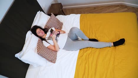 la joven está acostada en la cama de su dormitorio y navega por la página de las redes sociales con su teléfono inteligente, tomada desde arriba, con aspecto serio