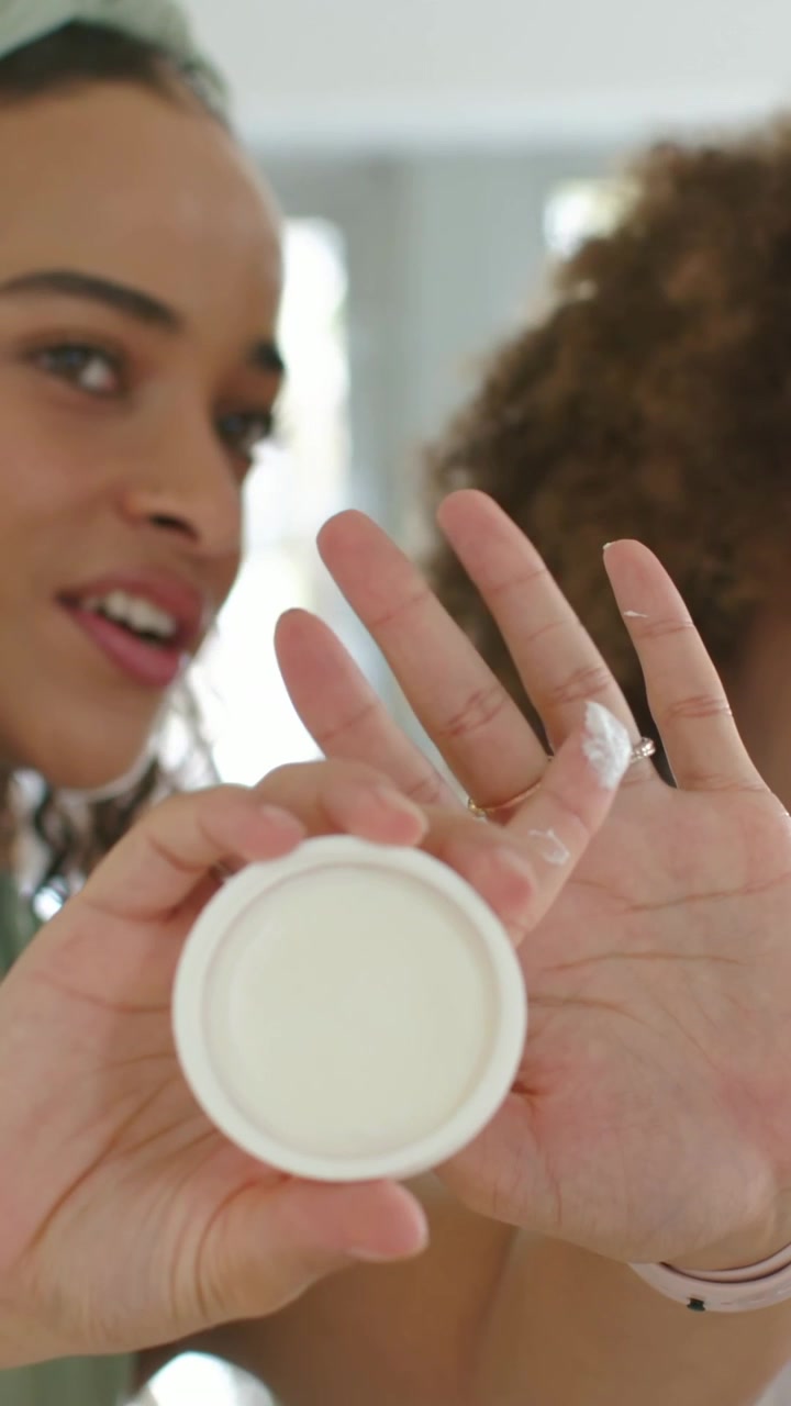 Vídeo vertical: Mulher com blusa verde virando o pote e aplicando creme na mão em casa para mostrar a textura.