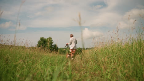 woman in white shorts walks ahead of her two dogs as they follow in grassy field under scattered sky, she holds leash and bag while leading pets on peaceful outdoor walk surrounded by green