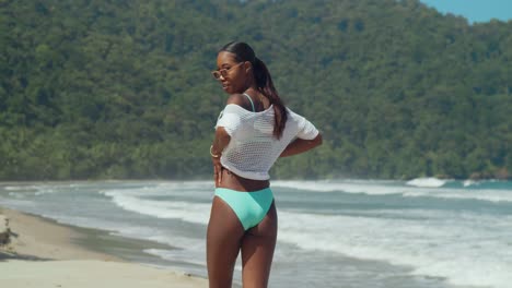 a girl in a breen bikini and netted top walks on a white sand beach with ocean waves in the background