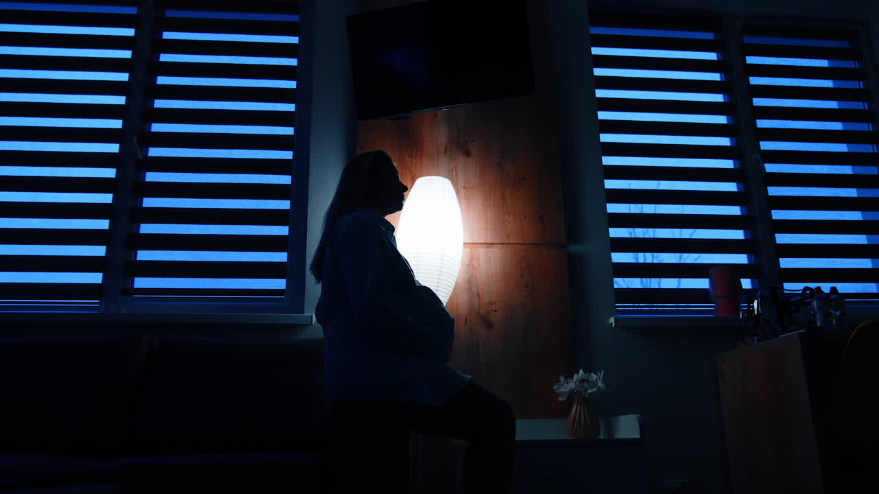 Calm long-haired pregnant woman sits on a sofa handrail in dark room. Future mom strokes her belly with love. Low angle view.