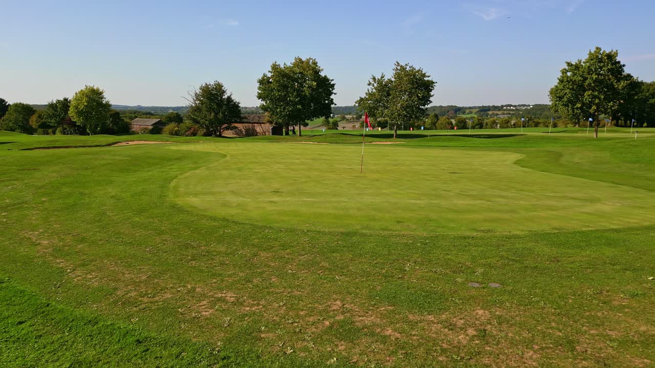 Wide shot pulling out from the golf green, with a centered red pin flag and close-cut grass surrounded by gentle slopes and mature trees, under clear summer skies