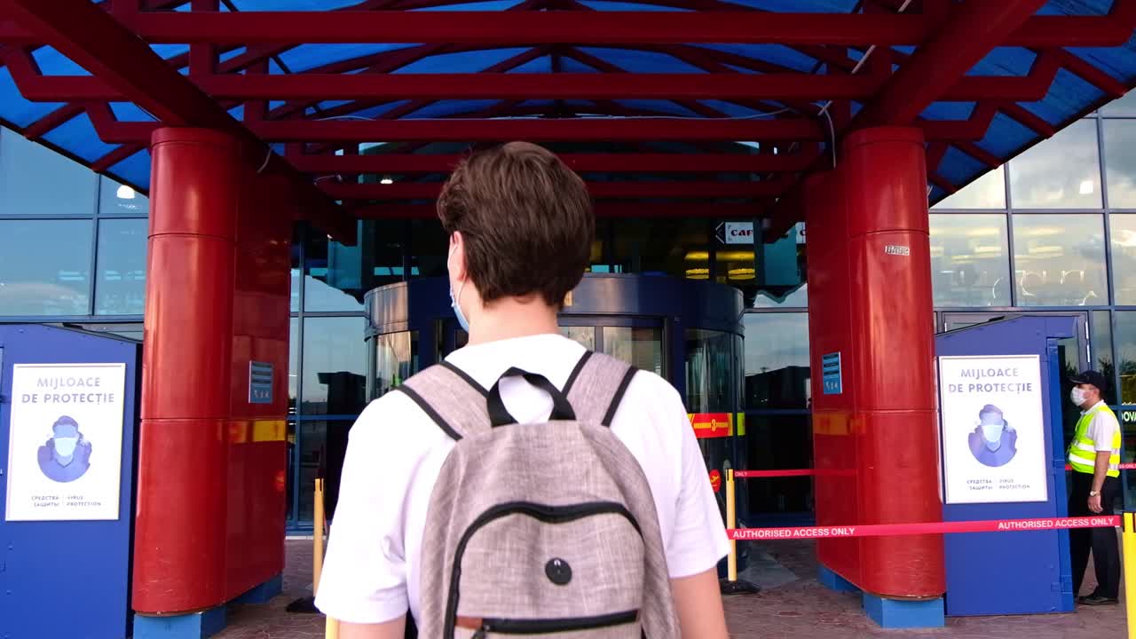CHISINAU, MOLDOVA - JUNE 19, 2020: Young man in white t-shirt, backpack and surgical mask walking to the airport entrance. Traveling during Corona Virus pandemic outbreak