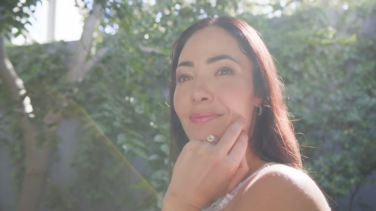 Câmera se afastando e luz do sol intensificando, mulher segurando o queixo, virando-se, fechando os olhos, sorrindo.