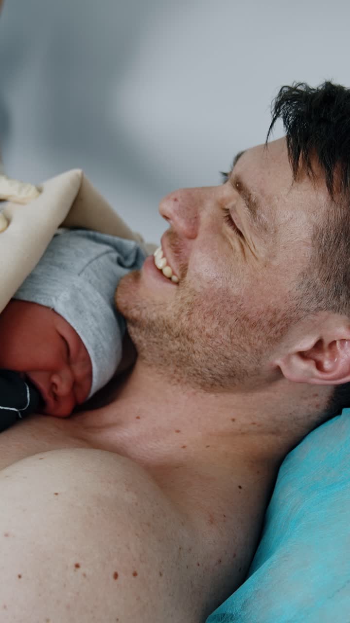 Il neonato viene posato sul petto nudo del padre. Il papà felice sorride. Un'infermiera aiuta a coprire il neonato. Video verticale.