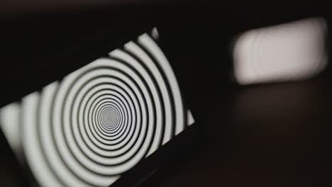 A-tablet-device-in-focus-and-a-mobile-phone-out-of-focus-in-a-dark-room,-both-displaying-a-hypnotic-spiral-pattern